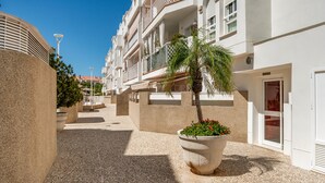 A courtyard with a palm tree, a large planter with flowers, and a building with balconies and a door labeled "POSTAL". - Apartment in Rota-Cádiz, new and modern near the beach. (Rota)