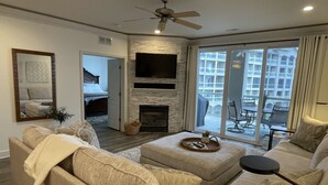 A modern living room with a fireplace, a ceiling fan, and a large window offering a view of a balcony. - Updated Lands End Upscale Condo - Sleeps 9 (Osage Beach)
