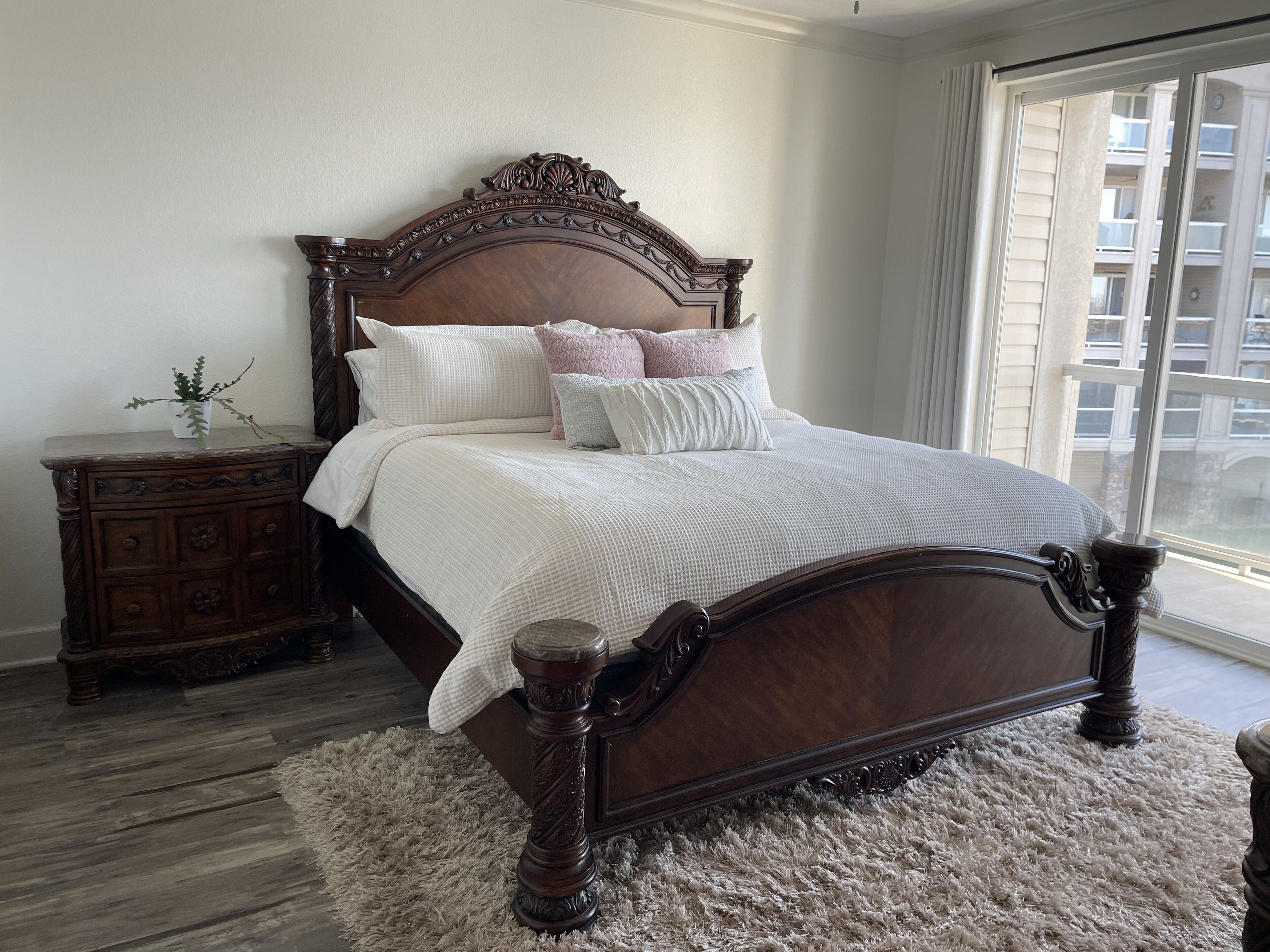 A bedroom with a wooden bed, a nightstand, a fluffy rug, and a window with a view of buildings.