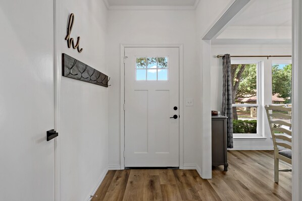 Un couloir avec une porte blanche, une petite table et une chaise près d’une fenêtre.