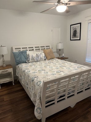 A bedroom with a bed, nightstands, a ceiling fan, a window with blinds, and a framed picture on the wall. - Winter Rates at Seize the Bay (Port St. Joe)