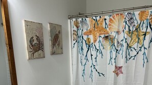 A bathroom with a white sink, a toilet, a shower curtain with sea shell designs, and a blue bath mat. - Winter Rates at Seize the Bay (Port St. Joe)
