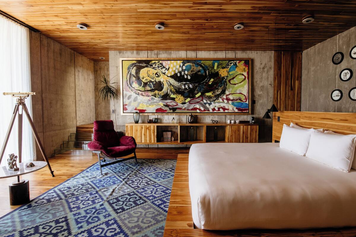 Una habitación con una cama grande, una silla roja, techo de madera y un cuadro abstracto y colorido en la pared.