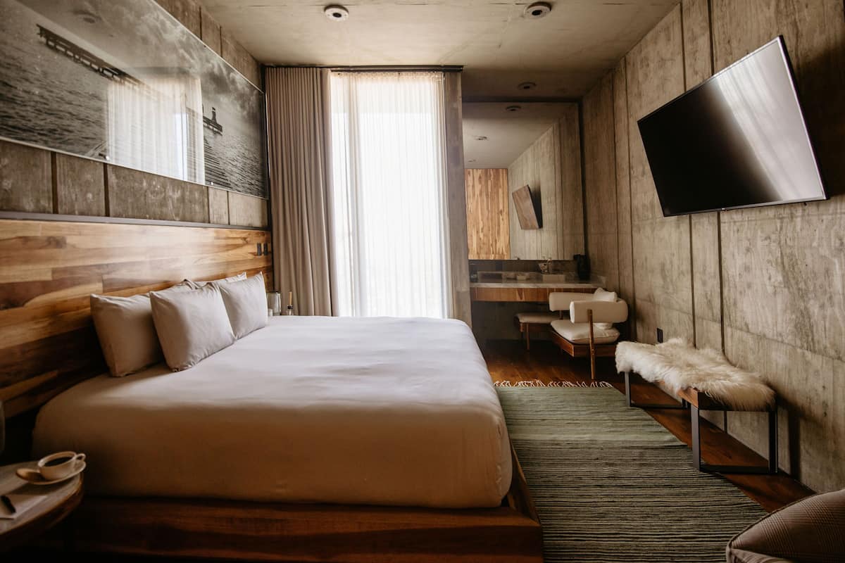 Habitación de hotel moderna con una cama grande, cabecera de madera, un televisor de pantalla plana, un pequeño espacio de descanso y un amplio ventanal con cortinas.