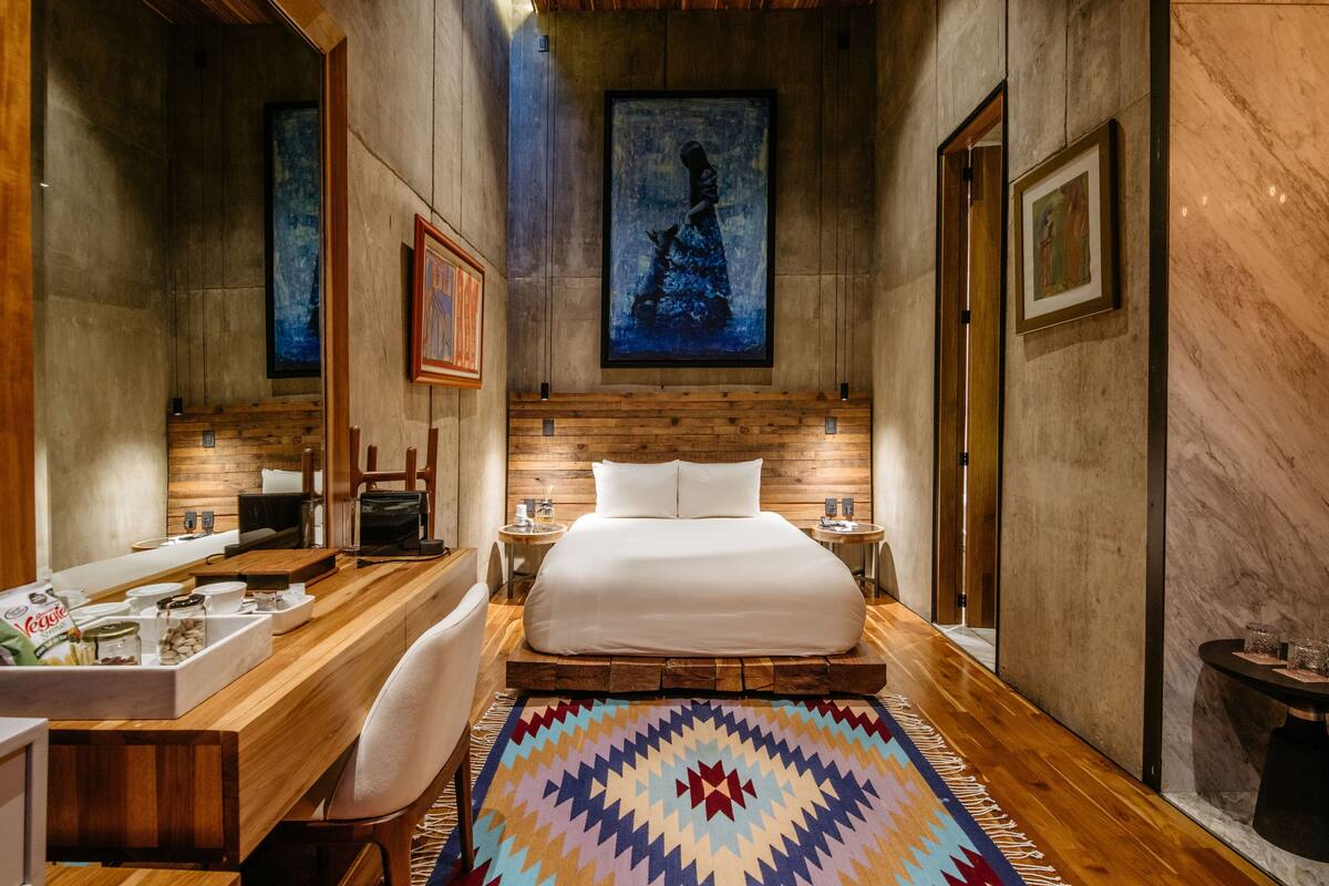 Habitación de hotel moderna con piso de madera, una cama grande, un escritorio de madera con una silla, un baño con espejo y obras de arte en las paredes.