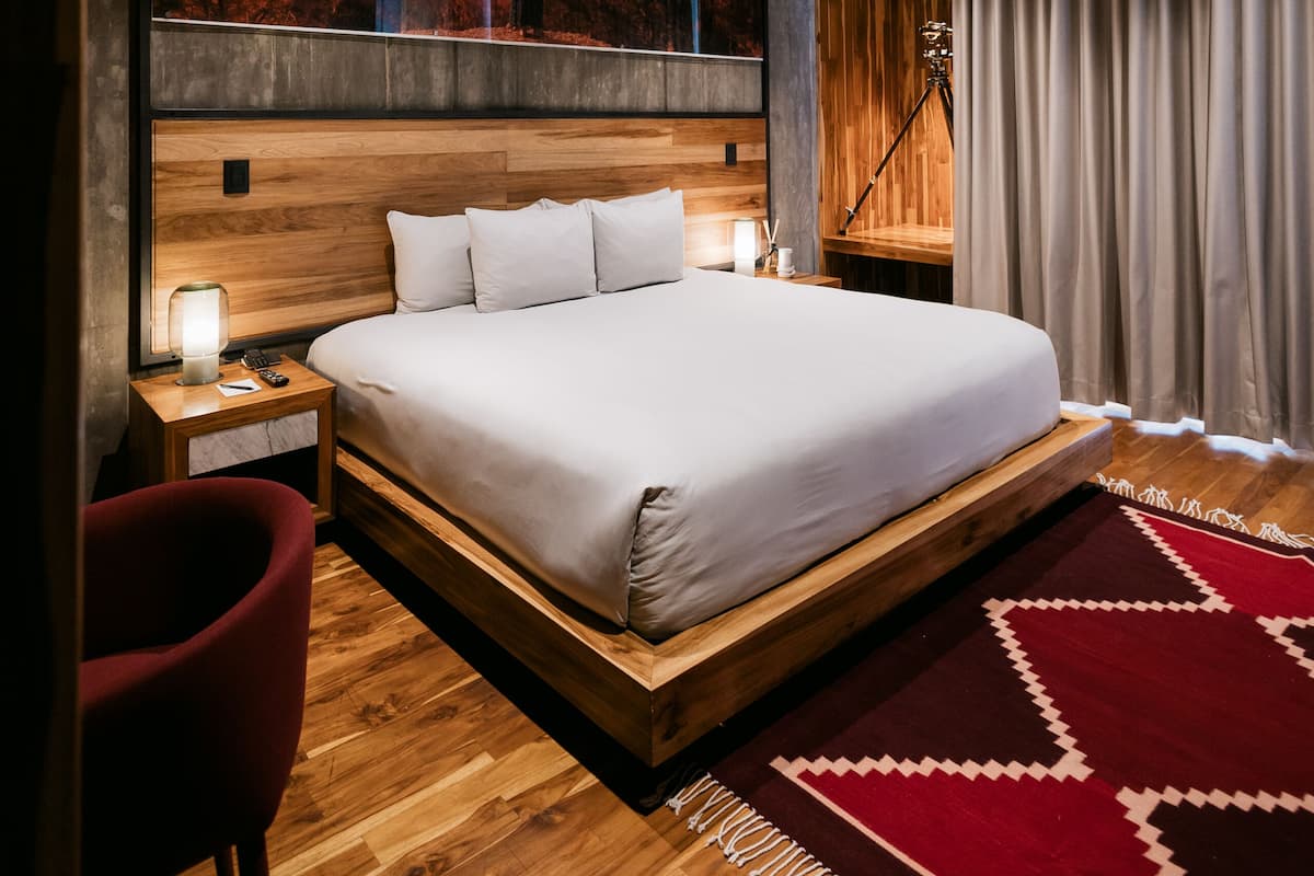 Un dormitorio moderno con cabecera de madera, una cama grande, un tapete estampado en rojo y beige, y una mesita de noche con lámpara.