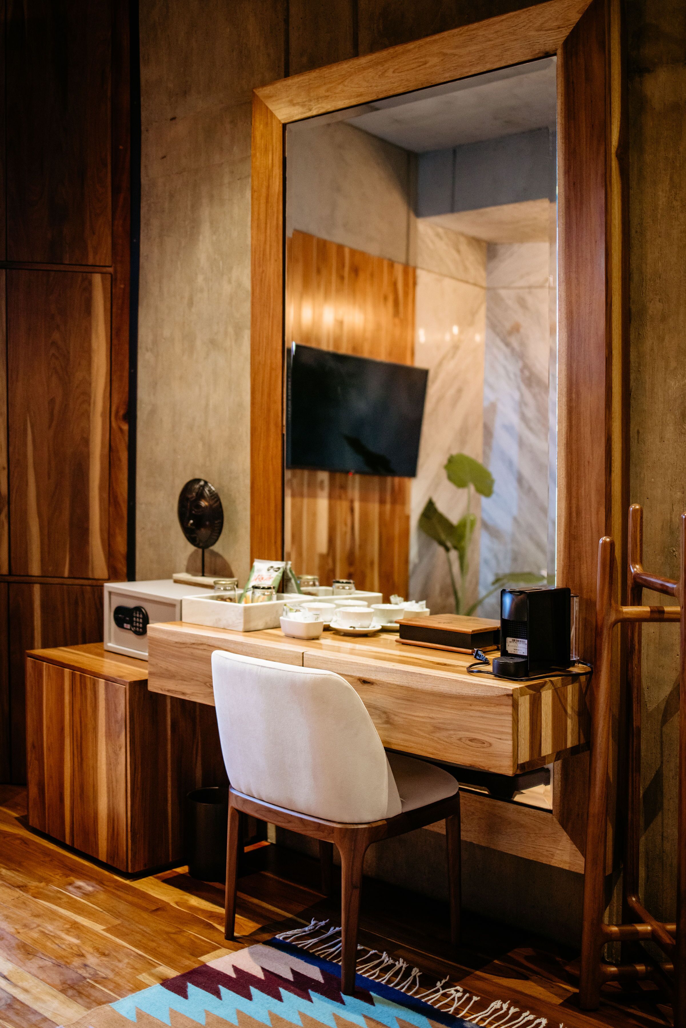 Un escritorio de madera con un espejo grande, una silla, una cafetera y un televisor montado en la pared.