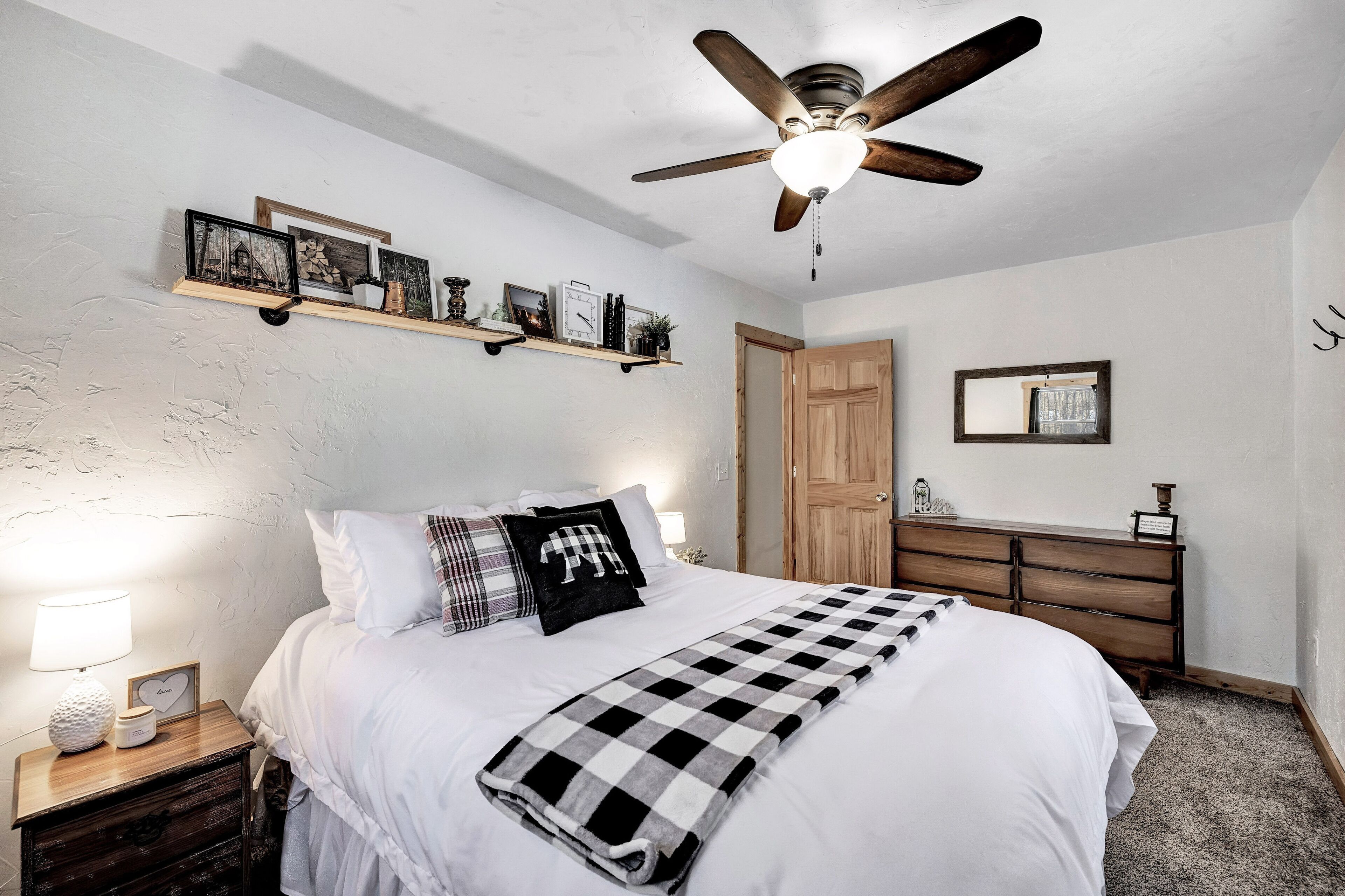 Quarto com cama, mesa de cabeceira, cômoda, ventilador de teto e prateleiras com quadros.