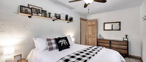 Quarto com cama, mesa de cabeceira, cômoda, ventilador de teto e prateleiras com quadros.