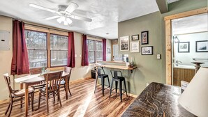 Un coin repas avec une table et des chaises en bois, un ventilateur de plafond et de grandes fenêtres ornées de rideaux rouges.