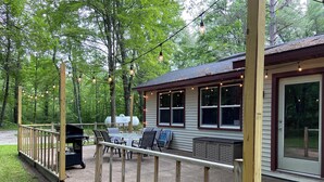 Une terrasse extérieure couverte, équipée d’un barbecue, d’une table à manger et de chaises, entourée d’une plateforme en bois et illuminée de guirlandes lumineuses.