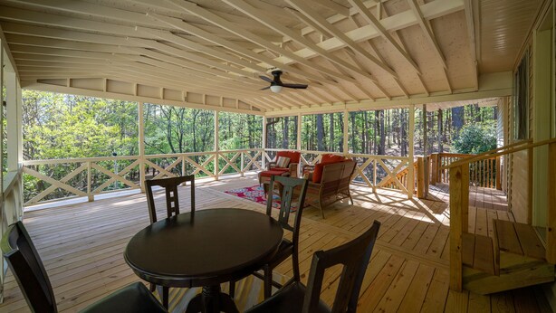 Terraza al aire libre techada con mesa y sillas, un ventilador de techo y vista al bosque.