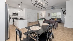 Une cuisine moderne avec des meubles blancs, un coin repas doté d’une table et de chaises noires, un canapé gris et un lustre géométrique original.