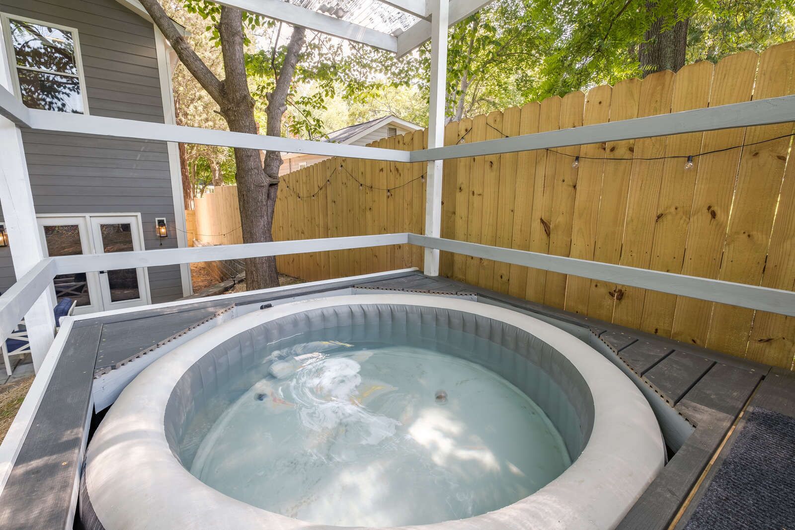 Un bain à remous dans un espace extérieur abrité, entouré d’une clôture en bois, avec une maison en arrière-plan.