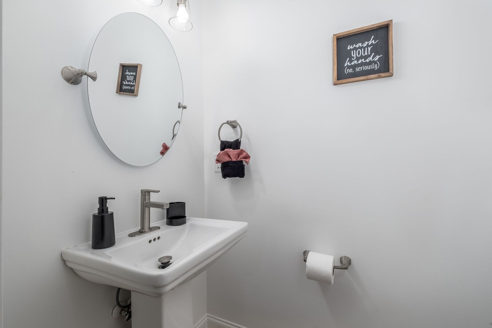Ein Badezimmer mit einem weißen Waschbecken, einem Spiegel, einem gerahmten Zitat an der Wand und einem Toilettenpapierhalter.