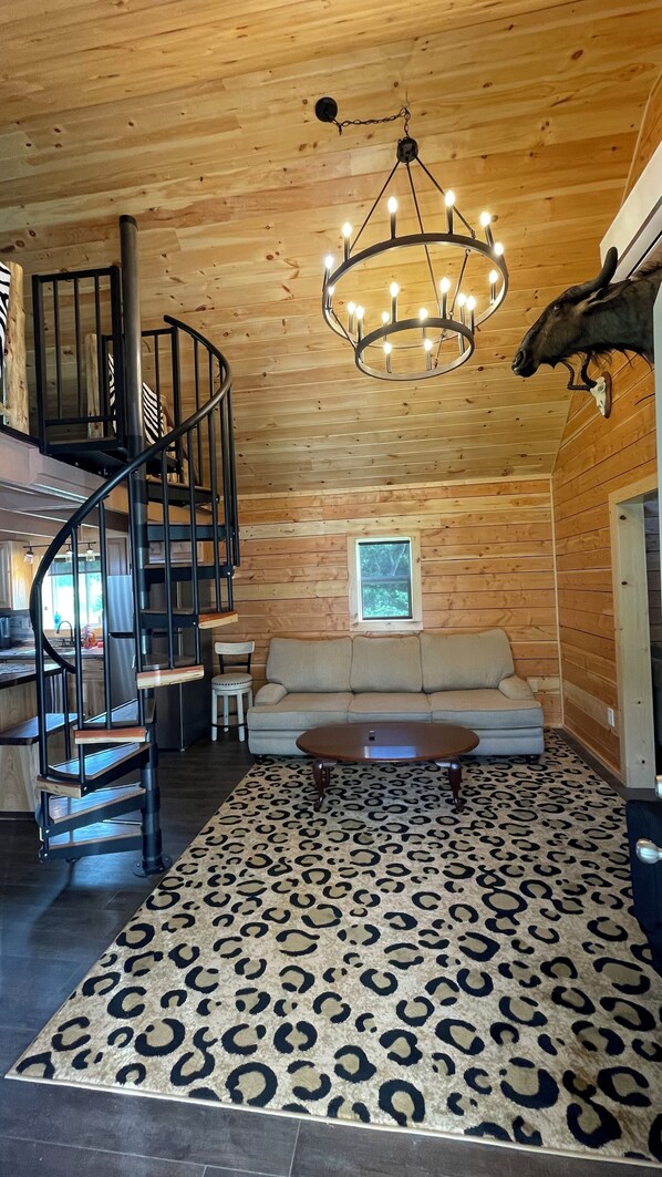 A wooden interior with a spiral staircase, a chandelier, a sofa, and a leopard print rug. - Renovated Cabin, with custom craftwork, in Picturesque Finger Lakes Wine Country (Branchport)