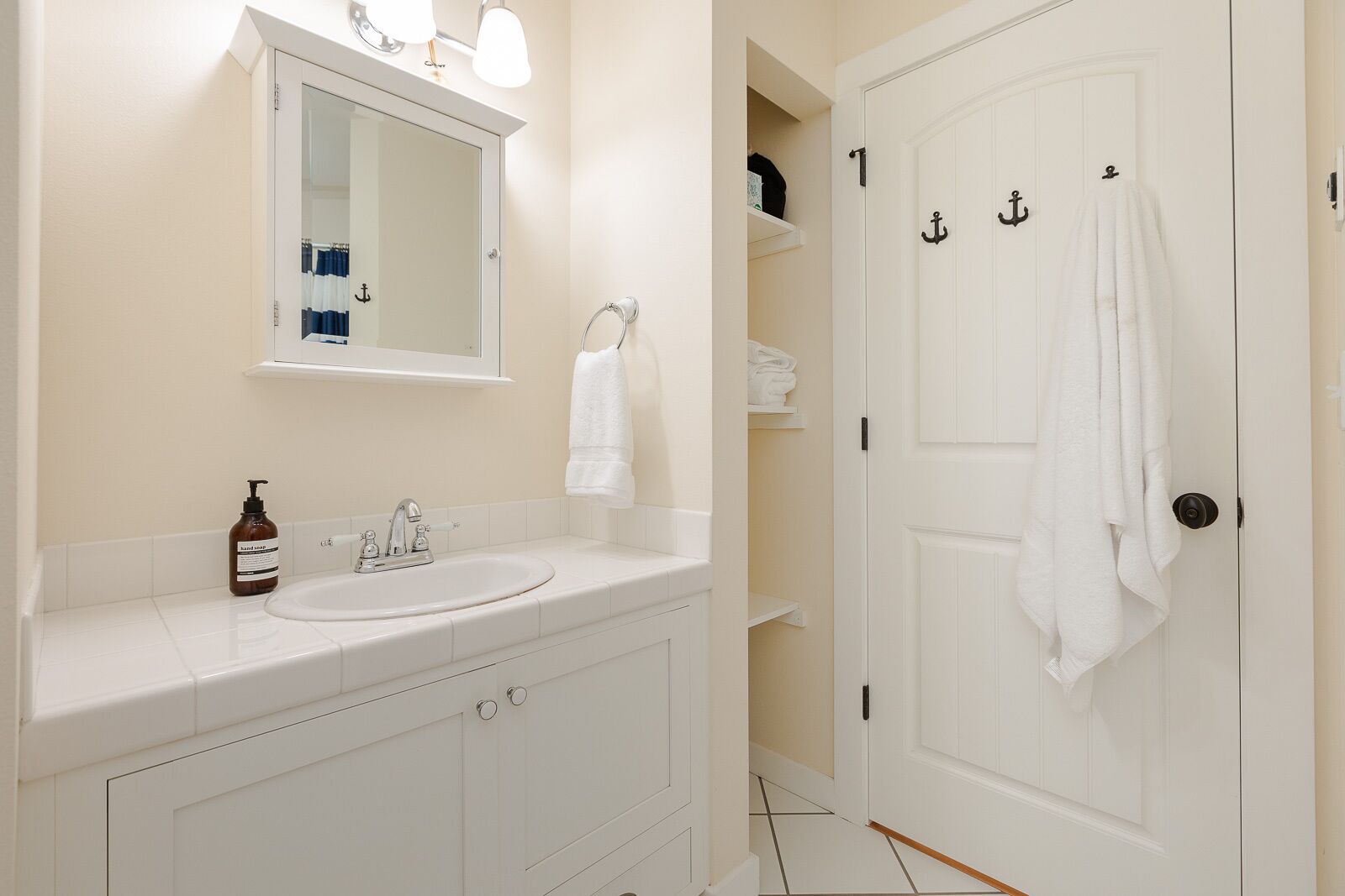 Une salle de bain avec un lavabo blanc, un miroir, un porte-serviettes et une porte dont les poignées sont en forme d’ancre.