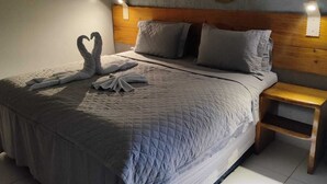 Una cama bien hecha con una colcha acolchada, dos almohadas y una disposición de toallas en forma de corazón. Se observa un cabecero de madera, una mesita de noche y un adorno en la pared sobre la cama.
