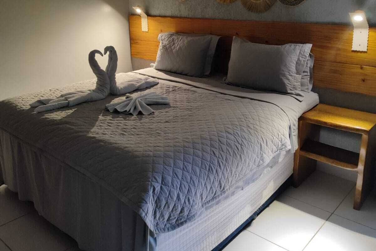 A neatly made bed with a quilted bedspread, two pillows, and a heart-shaped towel arrangement. There is a wooden headboard, a bedside table, and a wall decoration above the bed.