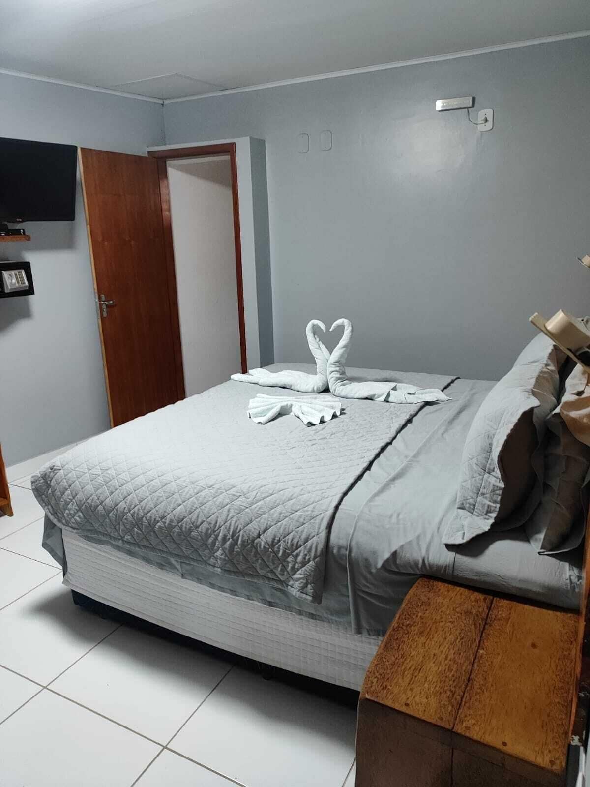 Un lit bien fait, avec une couette grise matelassée, une table de chevet en bois sur laquelle se trouve une lampe, et une décoration artistique réalisée avec des serviettes, posée sur le lit.
