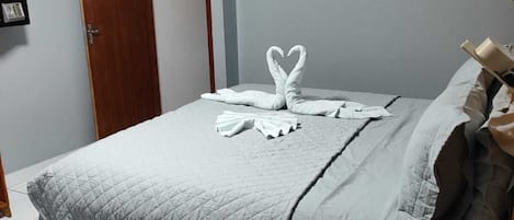A neatly made bed with a quilted grey duvet, a wooden nightstand with a lamp, and a towel art display on the bed.