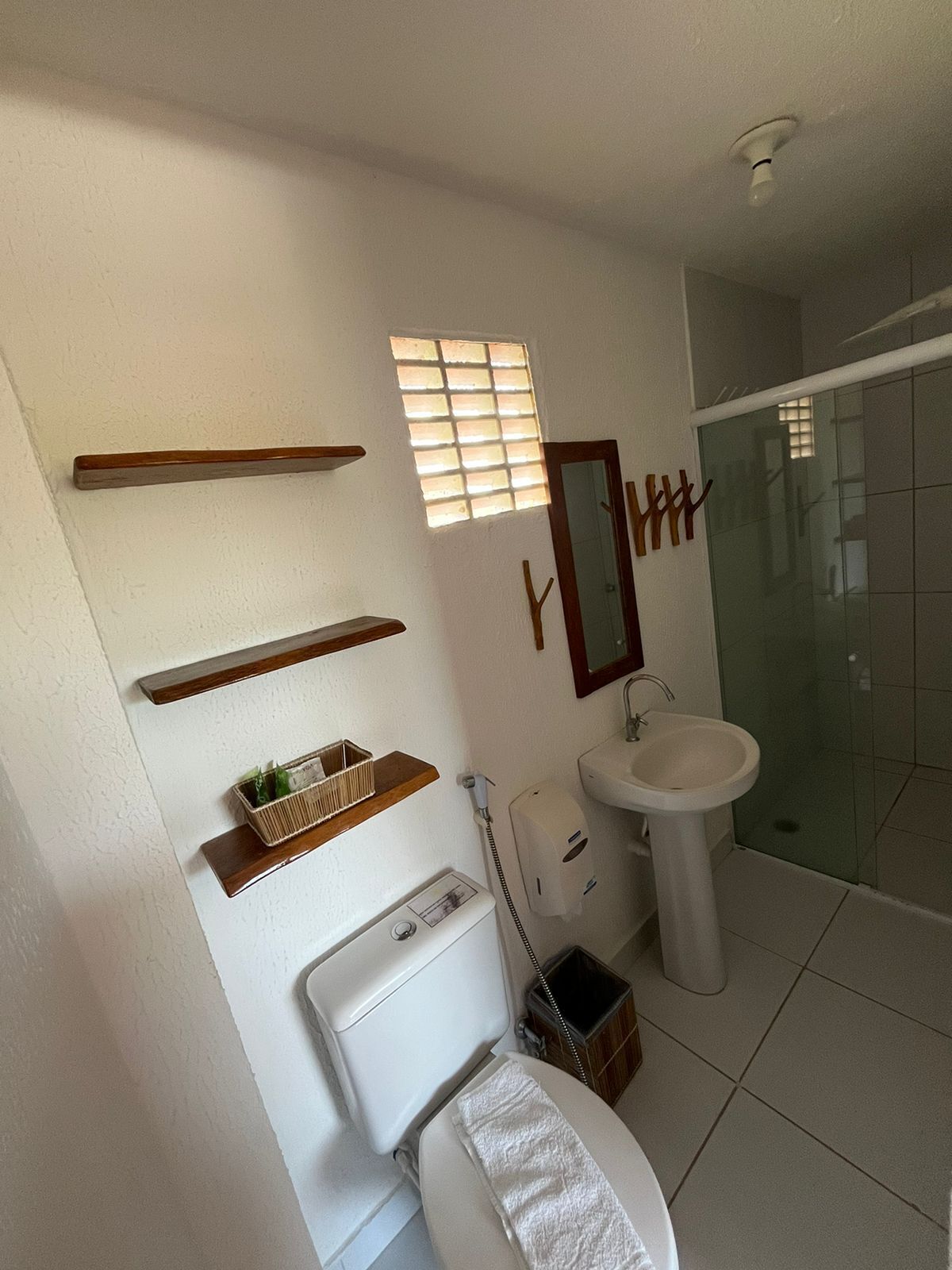 Un baño con inodoro, lavabo y ducha. Hay repisas de madera sobre el inodoro y una pequeña ventana sobre el lavabo.