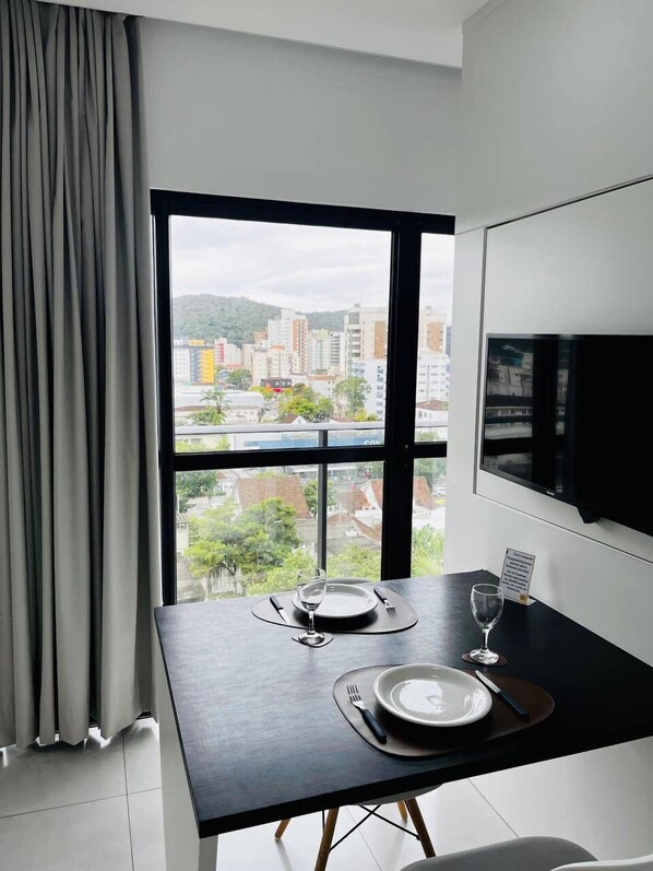 A modern dining area with a table set for two, a large window with a city view, and a flat-screen TV mounted on the wall.