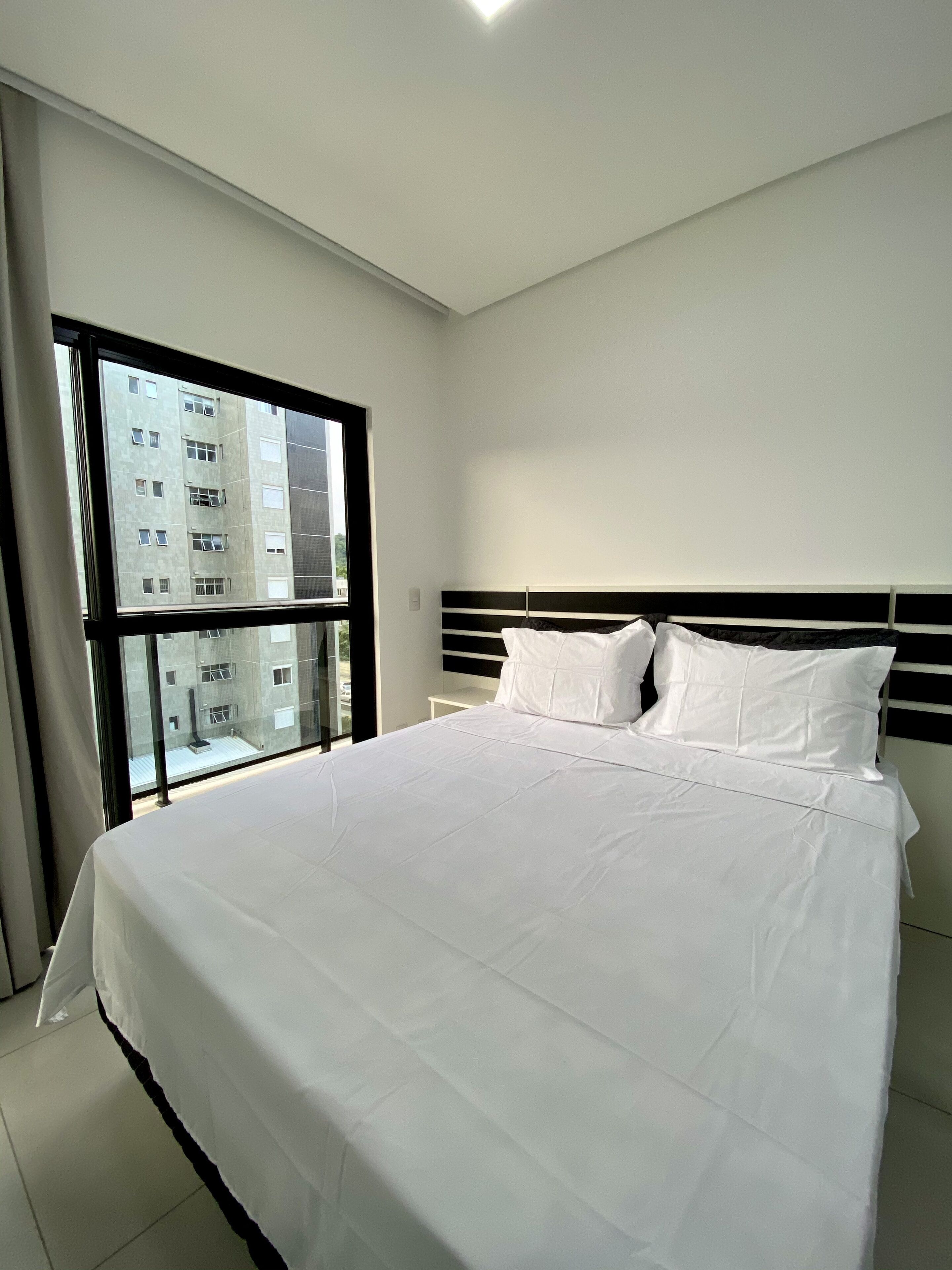 A modern bedroom with a large bed, a black and white headboard, and a view of a building through a large window.