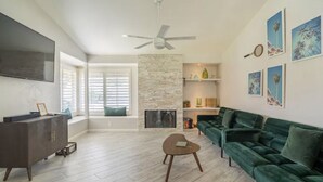A spacious living room with a large green sofa, a flat-screen TV, a stone fireplace, and a ceiling fan. - 3bed 2bath House.  Private Pool and Spa. Close to Acrisure Center  (Indian Wells)