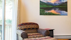 A cozy room with a wooden ceiling, a large window, a patterned armchair, a rug with animal prints, and a scenic painting on the wall. - Mountain Getaway and Family Retreat (Woodland Park)
