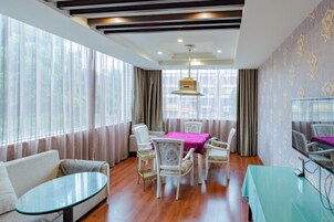 Habitación de hotel con zona de comedor, sofá, televisor y ventanas amplias.