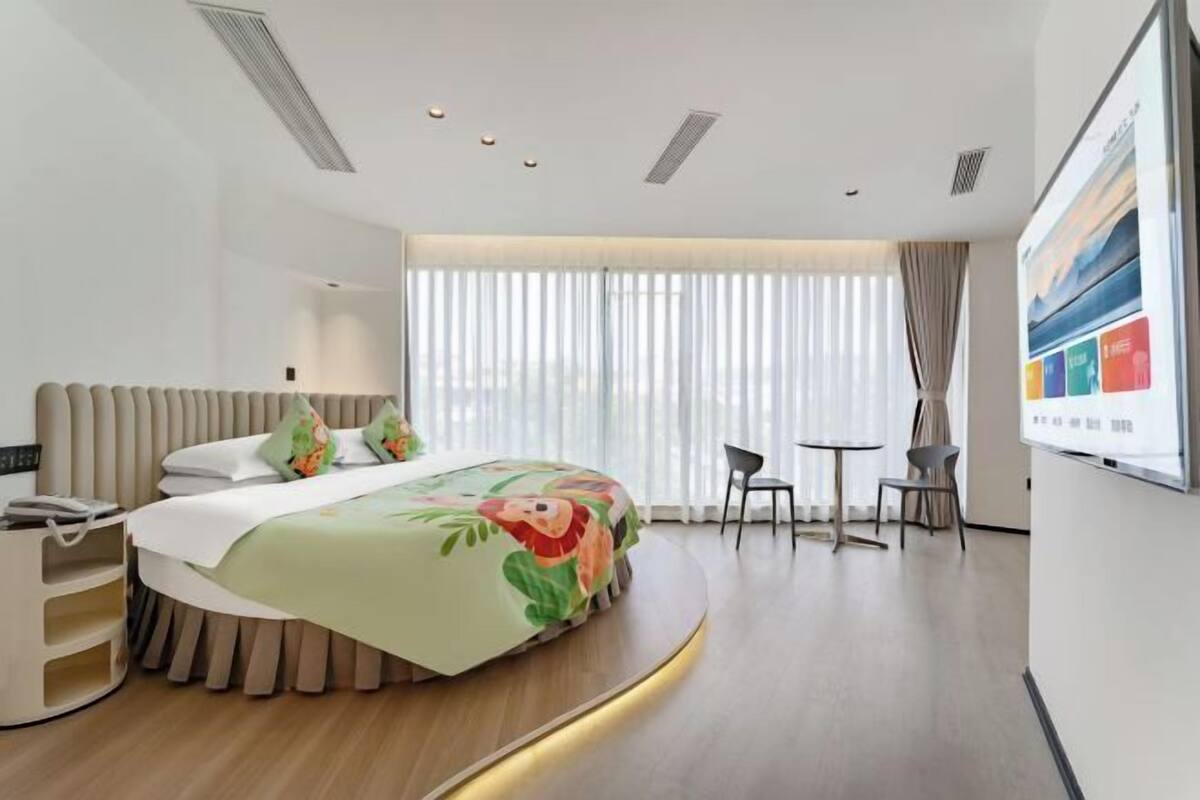 Quarto de hotel moderno com uma cama arredondada, escrivaninha, cadeira e uma janela grande com cortinas.