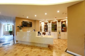 Zona de recepción de un hotel, con mostrador de recepción, un televisor montado encima y una zona de bar con estanterías llenas de botellas.