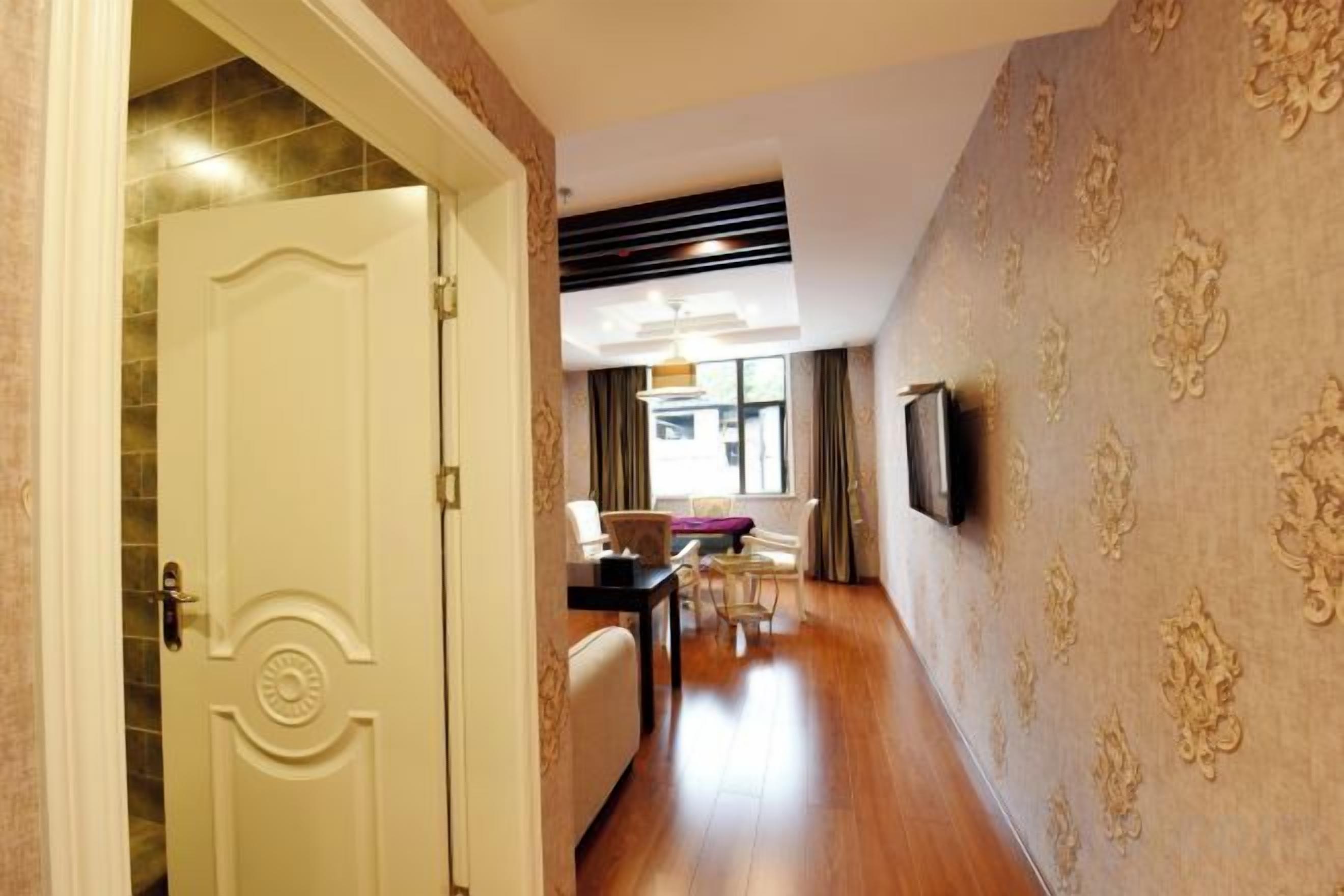 A hotel hallway with a closed door, a dining area with a table and chairs, and a wall-mounted television.