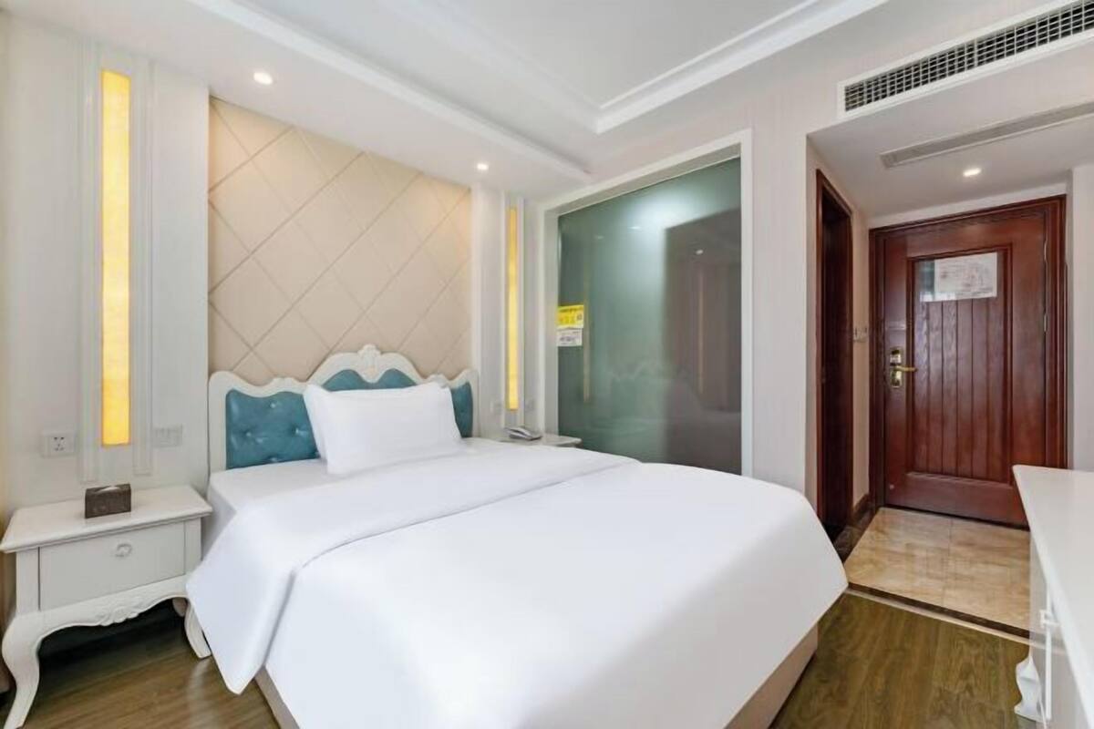 Quarto de hotel com uma cama grande, uma mesa de cabeceira, uma mesinha de noite, uma porta de madeira e um espaço para o banheiro.