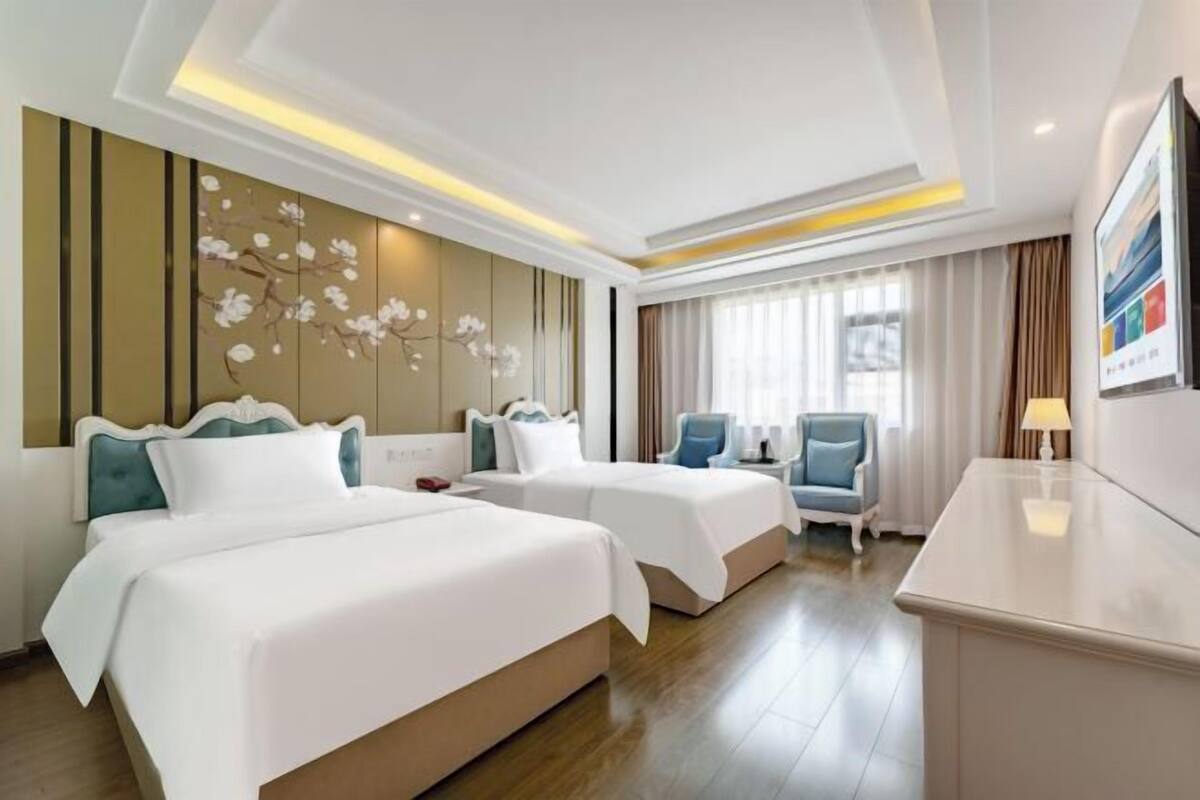 Quarto de hotel com duas camas, uma escrivaninha, uma cadeira, uma televisão e um painel decorativo na parede.