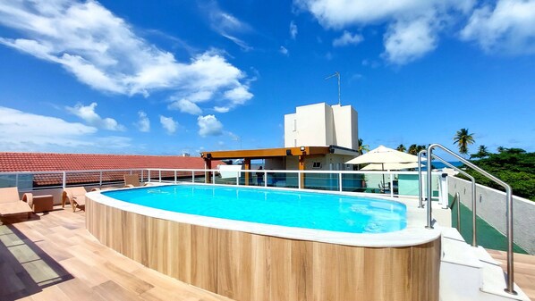 A rooftop pool with a wooden deck, lounge chairs, and a view of the ocean and palm trees.