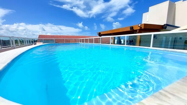 A clear blue swimming pool with a white deck, surrounded by a glass railing and a building with a red-tiled roof in the background. - Residencial Engenheiro Paulo Borba (Ipojuca)