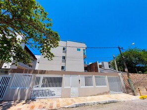 A multi-story residential building with a gated entrance, a clear blue sky, and a tree in the foreground.