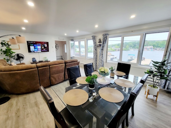 A modern living room with a brown sofa, a dining table with placemats, a TV mounted on the wall, and large windows with a view of a body of water.