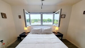 A bedroom with a large bed, two bedside tables with lamps, and a view of a body of water through two open windows.