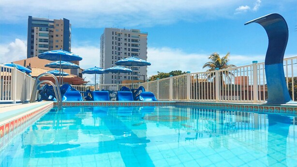 A swimming pool with blue umbrellas, a white fence, and tall buildings in the background. - Pousada Raio de Sol (Aracaju)