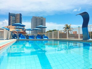 A swimming pool with blue umbrellas, a white fence, and tall buildings in the background.