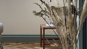 A room with a wooden floor, a window with a view, a vase with dried plants, and a wooden table.