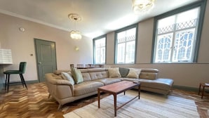 A spacious living room with a large L-shaped sectional sofa, a wooden coffee table, and a herringbone hardwood floor. There are large windows with decorative glass panes and a chandelier.