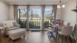 Uma sala de estar acolhedora com sofá, puf e área de jantar com cadeiras e mesa. Há uma varanda com vista para o exterior.