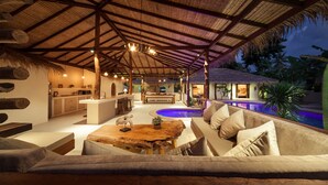 Un vaste espace de détente extérieur doté d’une piscine, de mobilier en bois et d’une structure à toit de chaume.