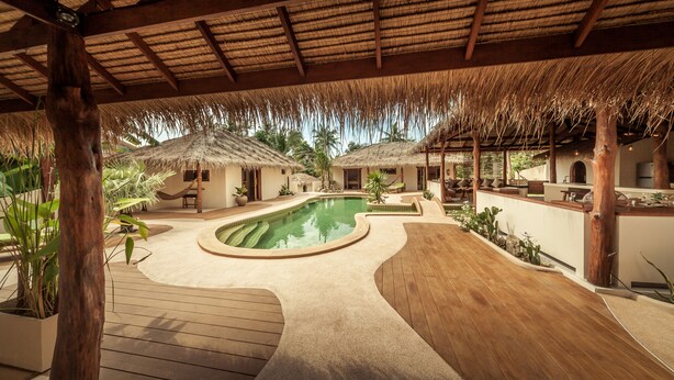 Un espace piscine doté d’une terrasse en bois, d’une structure à toit de chaume et d’une petite cascade décorative.