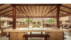 Un vaste espace extérieur aménagé en cuisine, doté d’un grand îlot central, de mobilier en bois et d’une structure à toit de chaume.