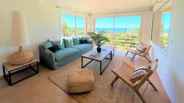 Amplia sala de estar con sofá, mesa de centro y sillas, ventanas grandes con vista al mar y un balcón.